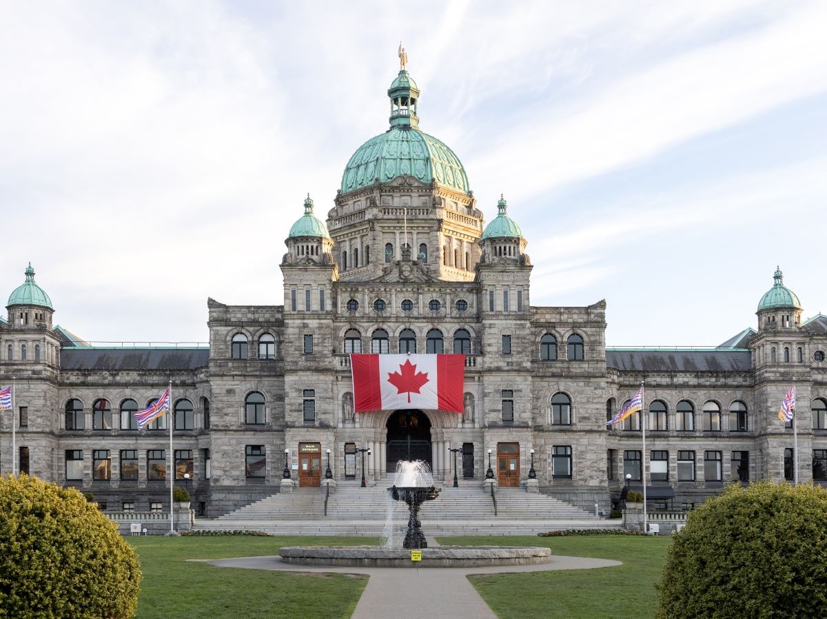 A large Edwardian stone edifice (the B.C. legislature) with a large red and white maple leaf flag hanging vertically in front of it.