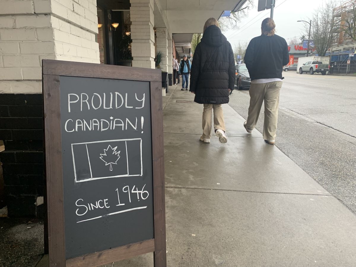 A hand-chalked sandwich board sign on a city street showing a Canadian flag and the words ‘Proudly Canadian since 1946’ with two people seen from behind walking by.