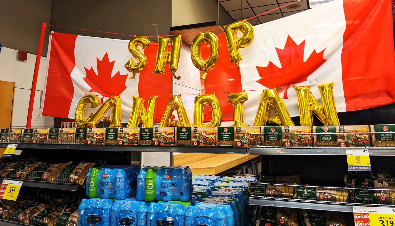 Gold balloon letters spell out ‘Shop Canadian’ in front of two Canadian flags hanging over store shelves with drinks and other products on display.