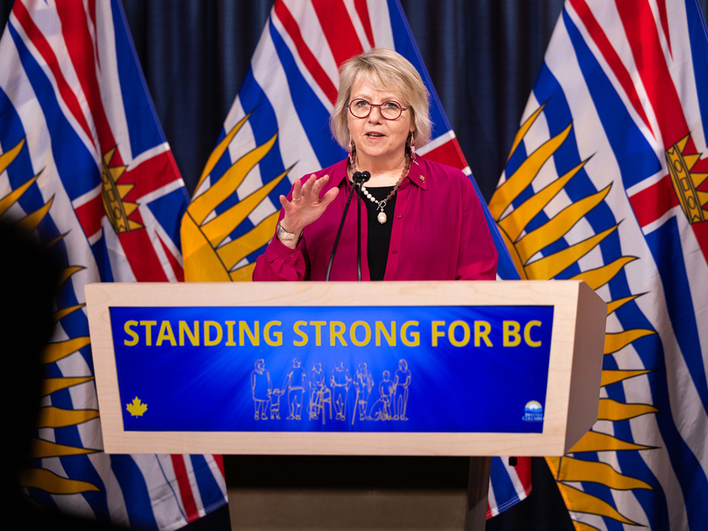 Dr. Bonnie Henry, a middle-aged woman with light skin tone and a blonde bob with bangs, wears red glasses, a black shirt, and a pink linen button-up shirt, and stands at a lectern that reads, ‘Standing Strong for BC.’