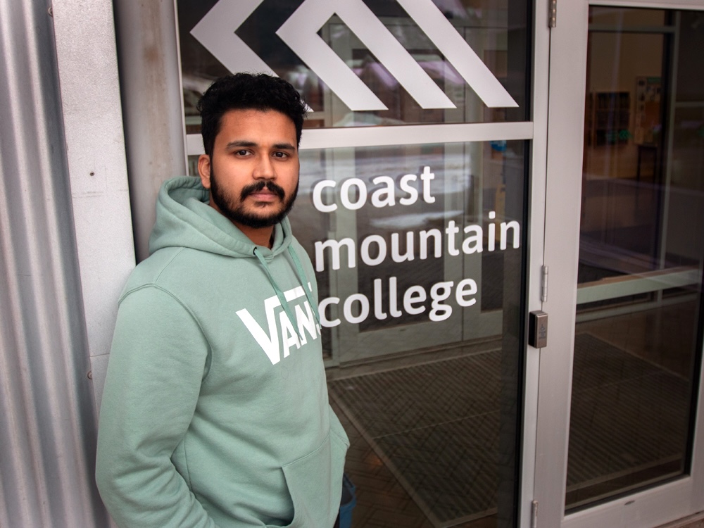 A man with medium-dark skin tone, short black hair and a beard, and wearing a hoodie, stands in front of glass doors with a logo that says ‘Coast Mountain College.’