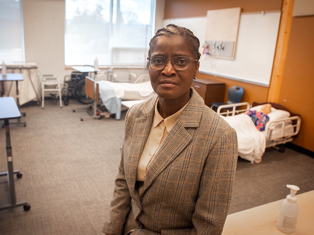 A woman with dark skin tone and braided hair, wearing glasses and business attire, sits in a room filled with hospital beds.