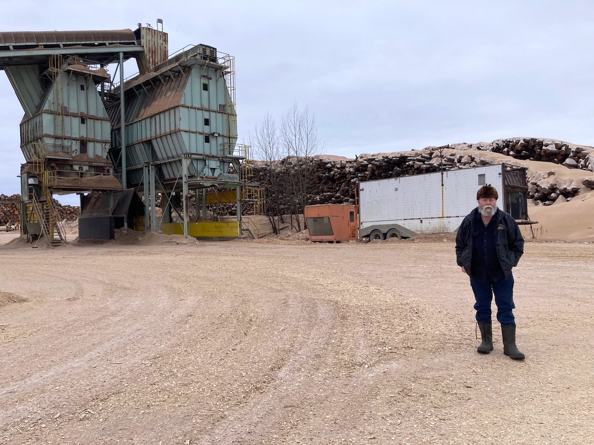An older man with light skin tone and a white beard wears a fur hat and stands a ways away from a large log pile and two large metal structures that are shaped a bit like extended stop signs.