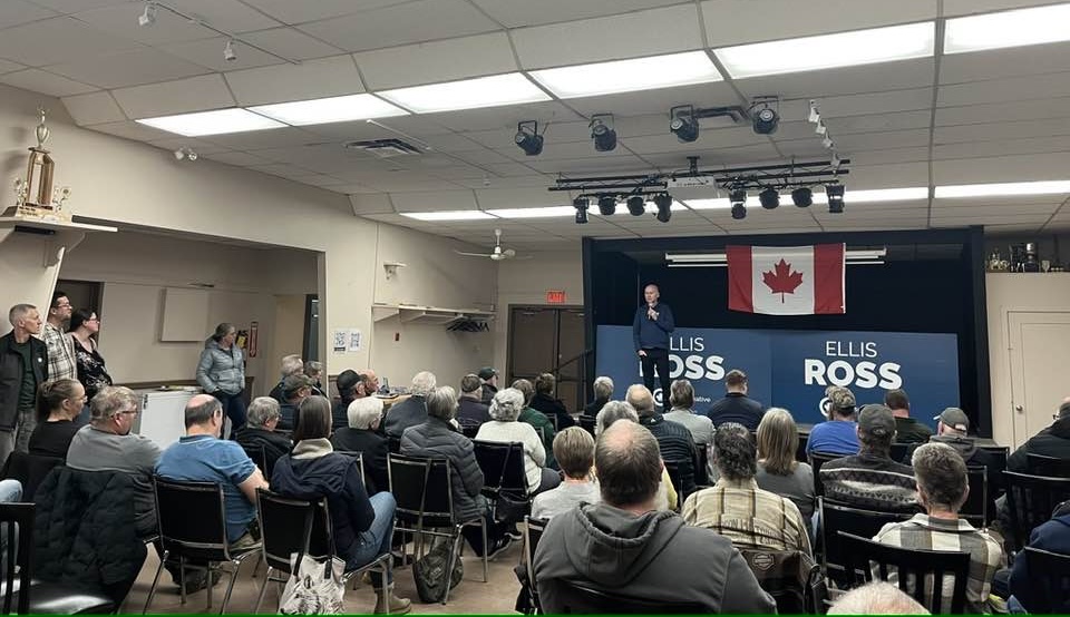 A man wearing dark clothing stands before an audience of several dozen people holding a microphone. Behind him are two signs that say ‘Ellis Ross.’