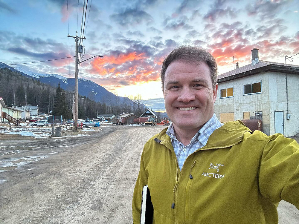 A man with fair skin tone and short hair, and extending his arm to take a selfie, stands in a residential neighbourhood in front of a mountain landscape and pink sunset.