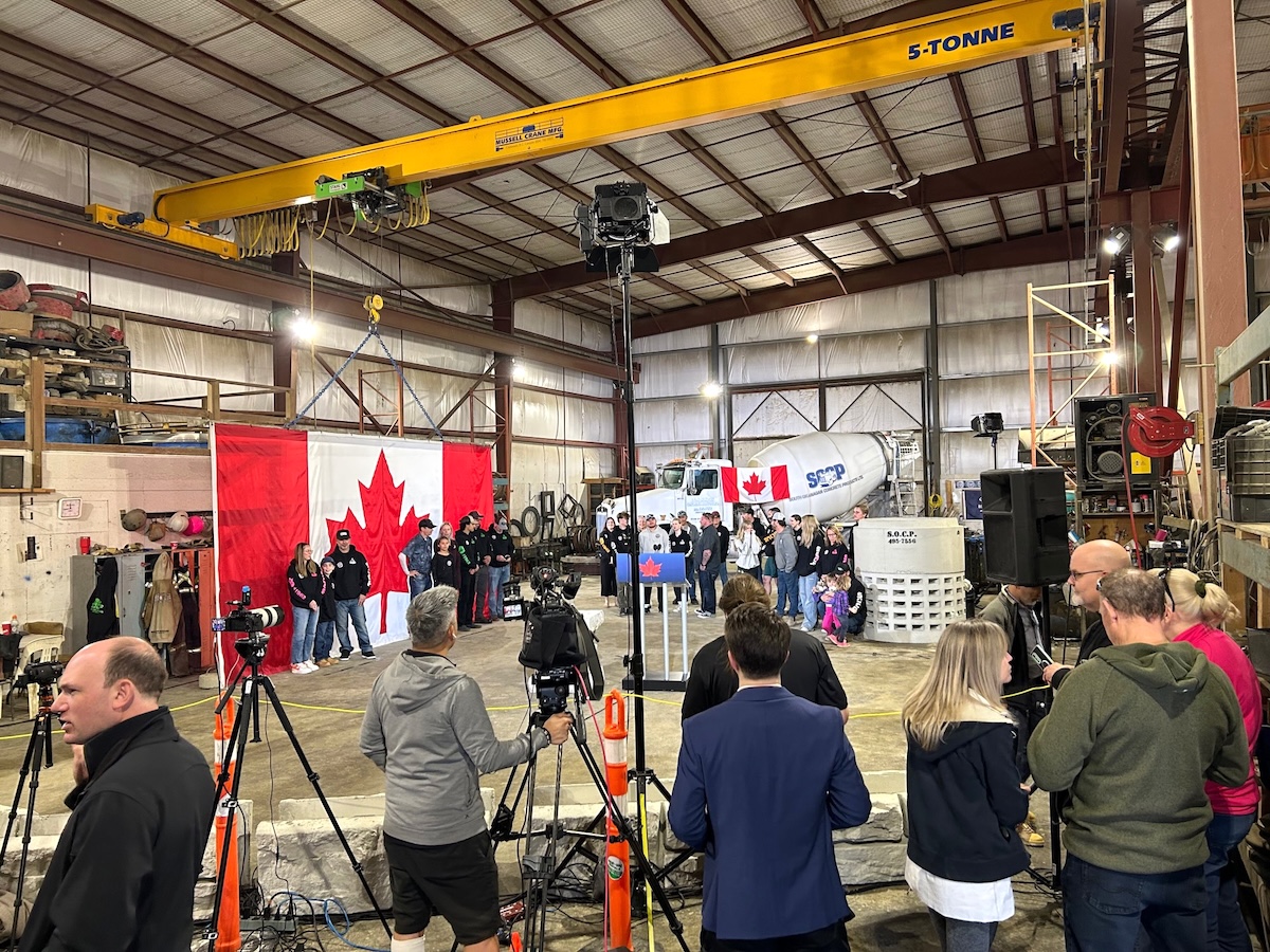 The photo shows the interior of a large industrial space. In the foreground is a handful of people, some with cameras on tripods. Beyond is a large Canadian flag with people standing in front of it.