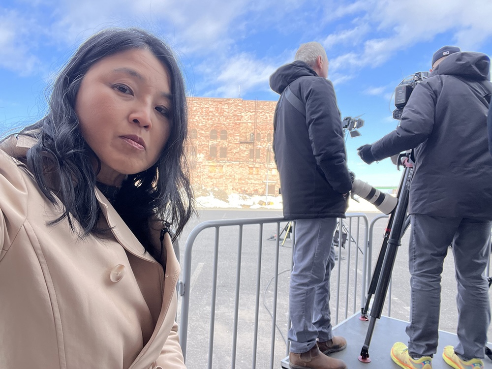 A woman with long dark hair and medium skin tone is in the foreground. Behind her there is a fence and two people holding camera equipment.