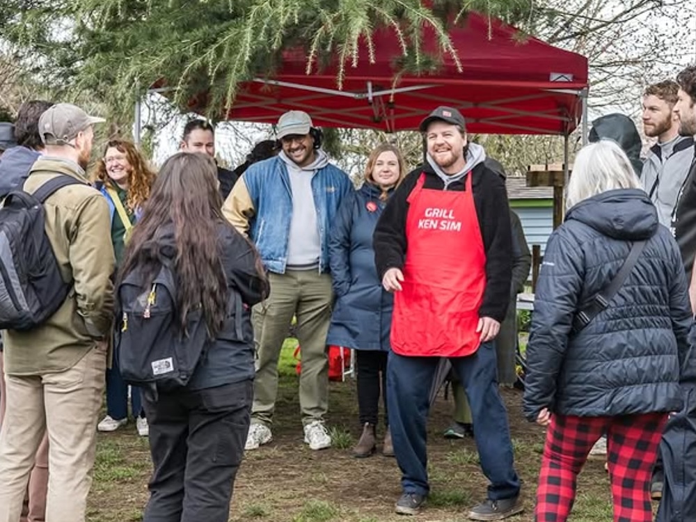 Sean Orr, a man with light skin and a beard stands among a group of smiling people. He is wearing a ball cap, a black jacket, jeans and a red apron that reads, ‘Grill Ken Sim.’
