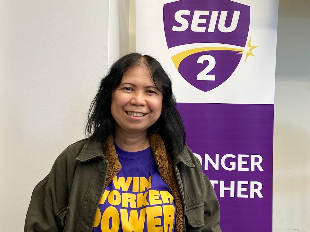 Emyll Garcia has dark wavy hair. She is smiling at the camera and wearing a purple T-shirt with yellow text that says ‘Win Worker Power.’ She is standing indoors next to a tall purple banner for the union SEIU Local 2.
