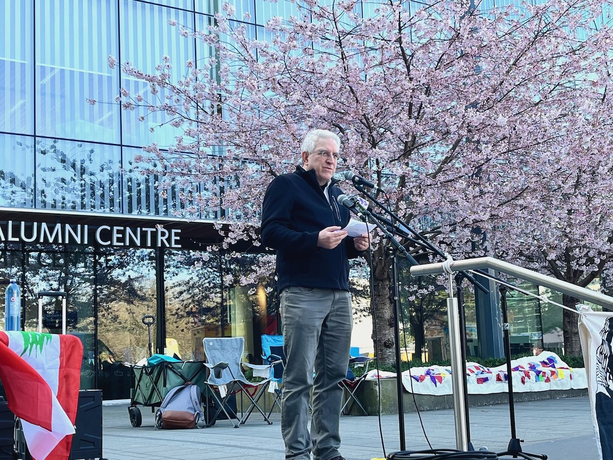 A middle-aged man with white hair, wearing a half-zip sweater, stands at a microphone. 