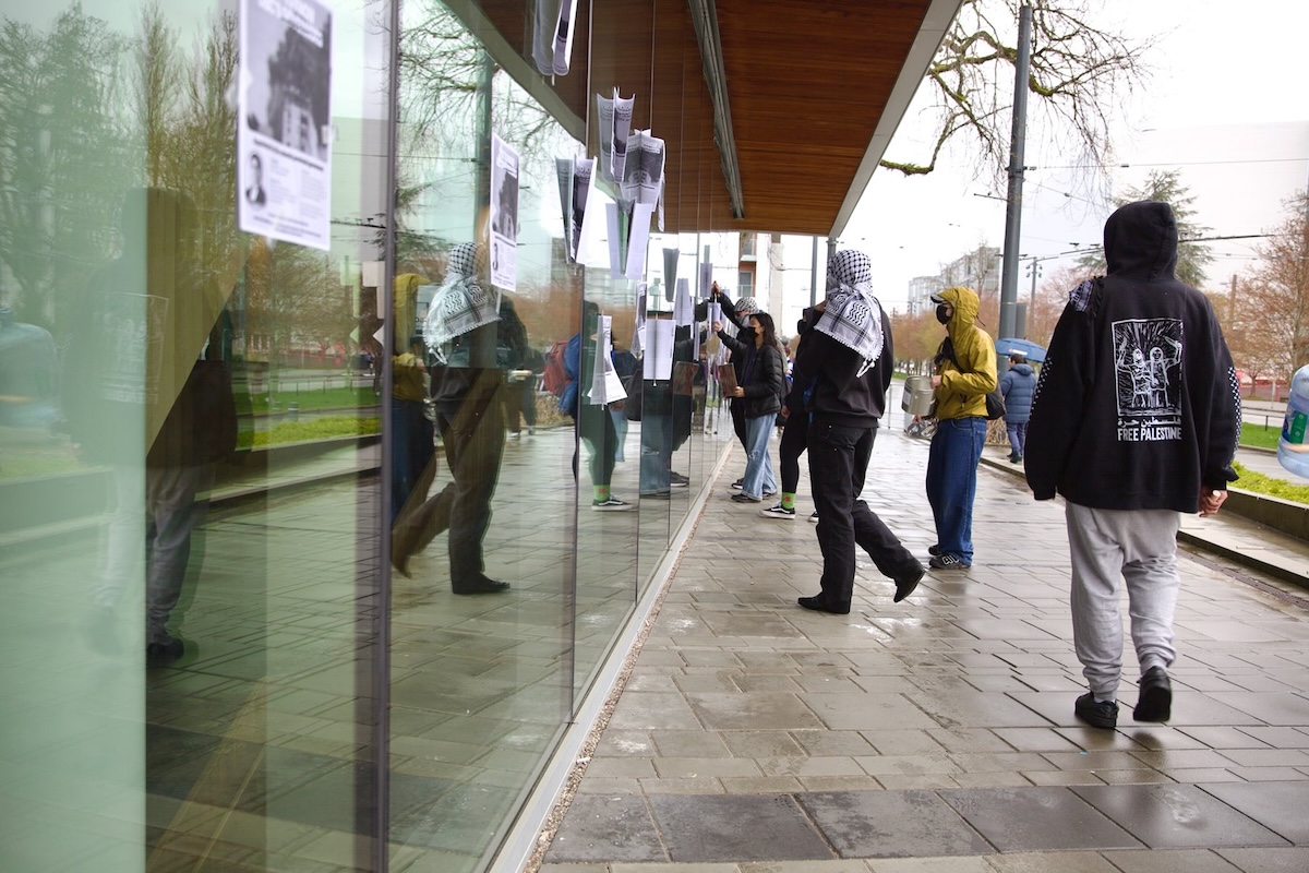 Students wearing kaffiyehs and face coverings tape letter-sized sheets of paper to glass windows. 
