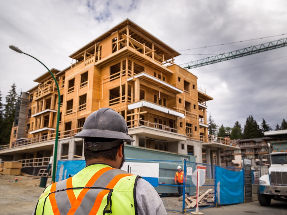 A construction worker wears a hi-vis vest and stands in front of a building under construction.