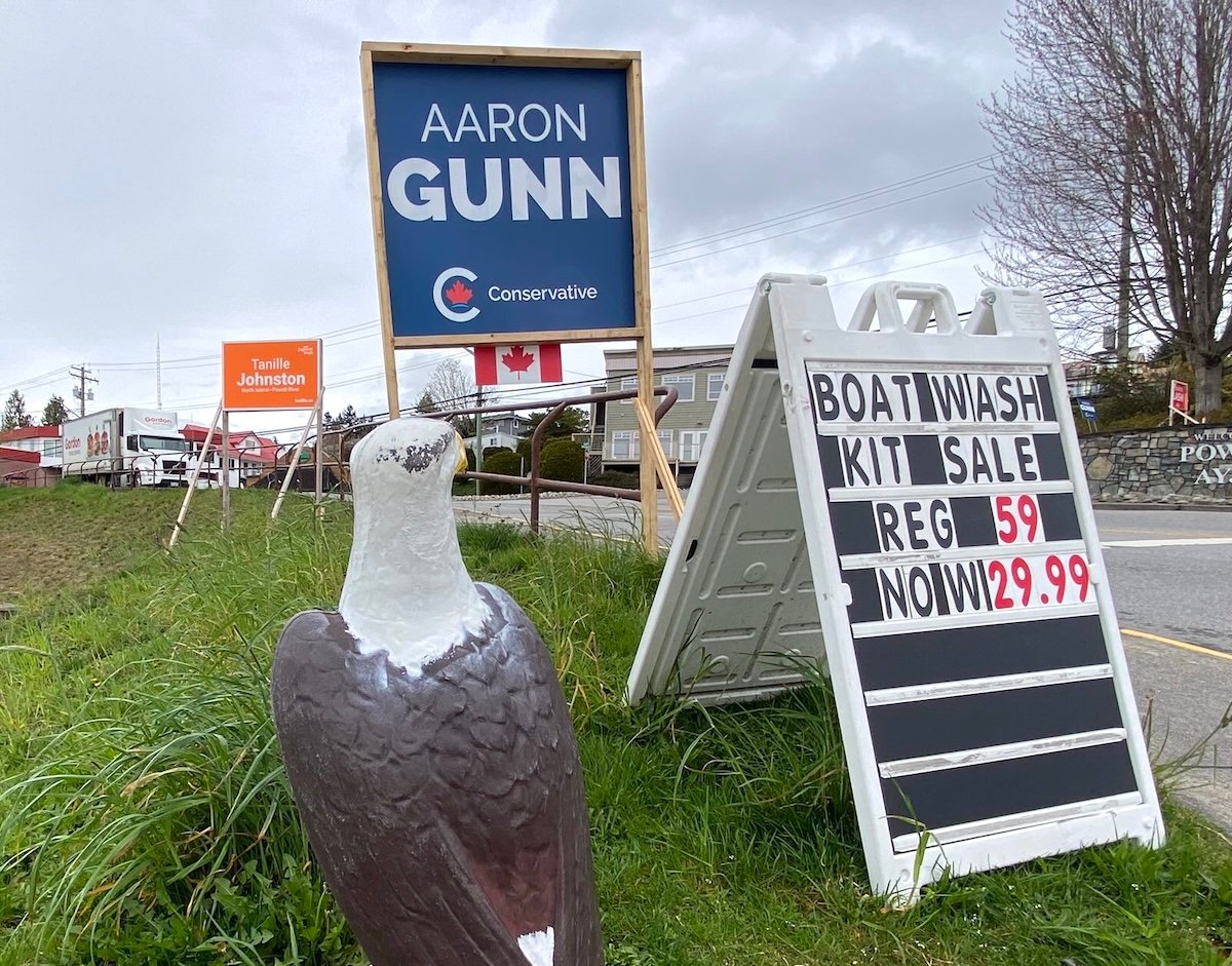 Electoral signs for Tanille Johnston and Aaron Gunn are posted at the corner of an intersection that also features a big plastic bald eagle and a sandwich board advertising a sale on boat wash kits. Across the street, a smaller sign for Jennifer Lash is just visible.