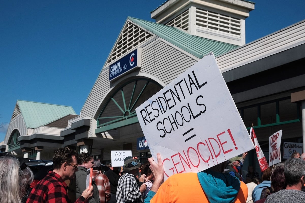 A sign on a building reads, ‘Gunn campaign HQ.’ People are gathered holding homemade signs. One reads, ‘Residential Schools = Genocide.’