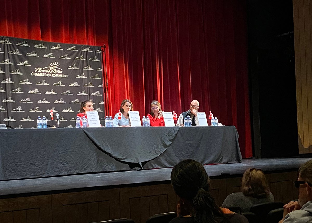 A spotlight illuminates the candidates onstage. They sit behind microphones at a long table. Behind them, a red curtain and a piece of black fabric with ‘Powell River Chamber of Commerce’ printed on it.