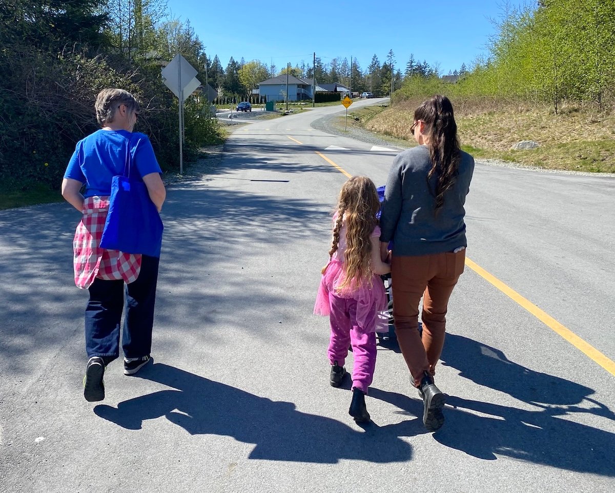 Two women and a child walk down a sunny street. The photo has been taken from behind them.