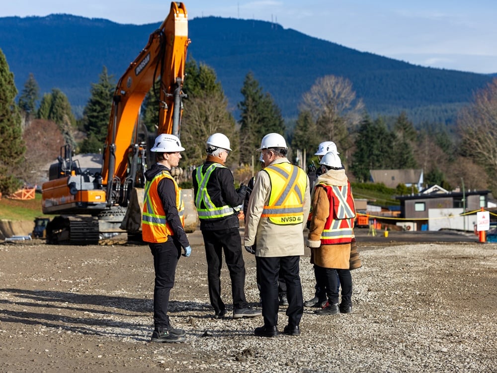 Five people wearing hard hats and high-visibility vests stand in front of construction equipment.