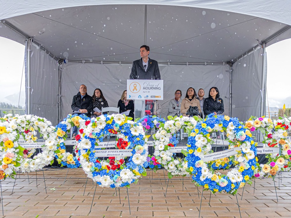 A man with short brown hair and pale skin tone stands at a lectern with a white sign that reads ‘Day of Mourning.’ He is wearing a button-down shirt under a long grey jacket. People are seated onstage behind him under a white tent. Colourful memorial floral wreaths are arranged on a beige tile floor in front of the stage.