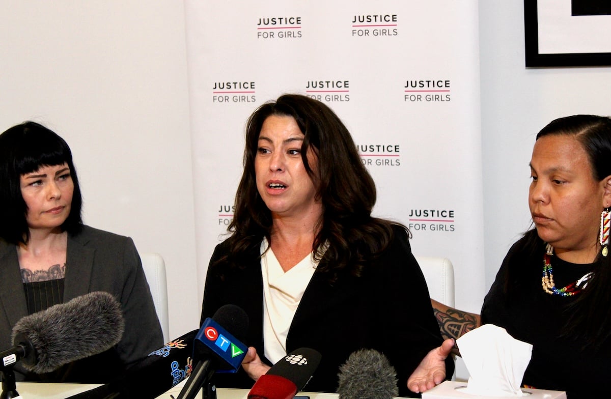 Three women with dark hair sit in front of a poster that reads ‘Justice for Girls’ with several press microphones lined up in front of them.