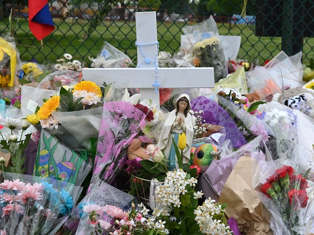 A white cross has been wrapped with a blue rosary. There is a figure of Mary praying. These religious symbols are surrounded by bouquets of flowers and some toys and stuffed animals.