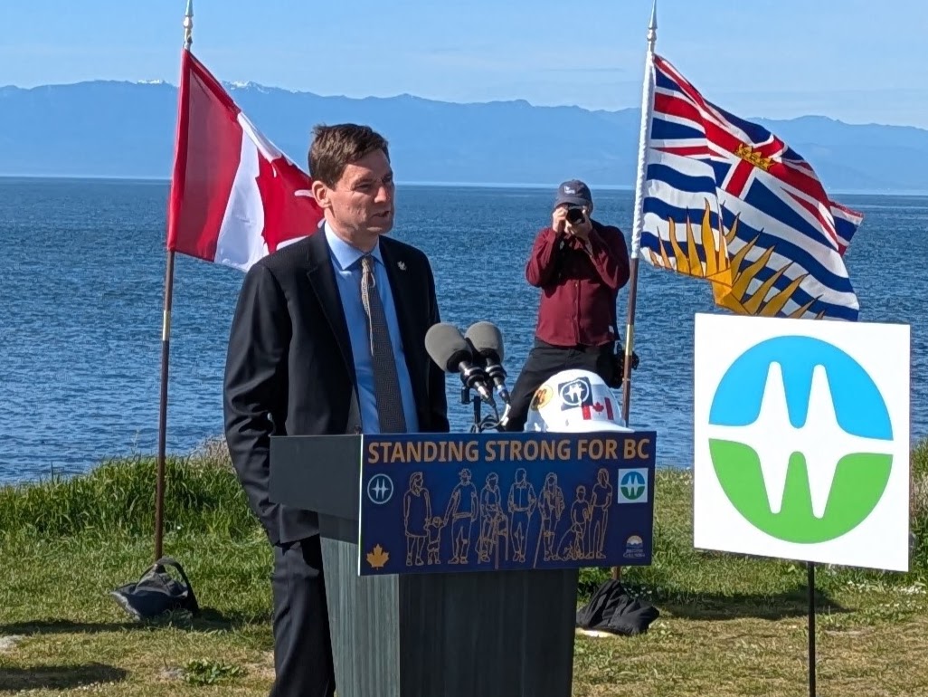 David Eby, a tall light-skinned man, stands outside at a podium that says, ‘Standing Strong for BC.’