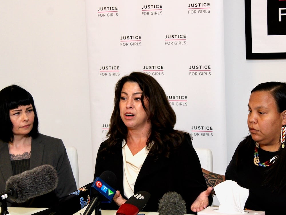 Three women with dark hair sit in front of a poster that reads ‘Justice for Girls’ with several press microphones lined up in front of them.