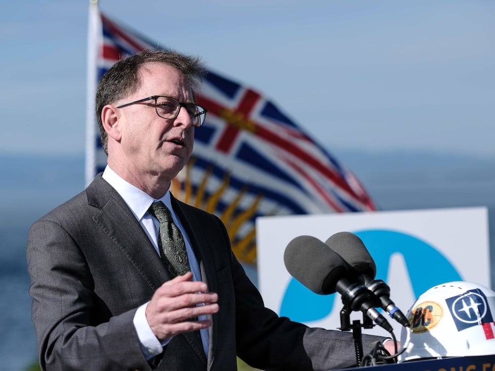 Adrian Dix, a 61-year-old light-skinned man with brown hair, stands outdoors at a lectern with an out-of-focus B.C. flag behind him.