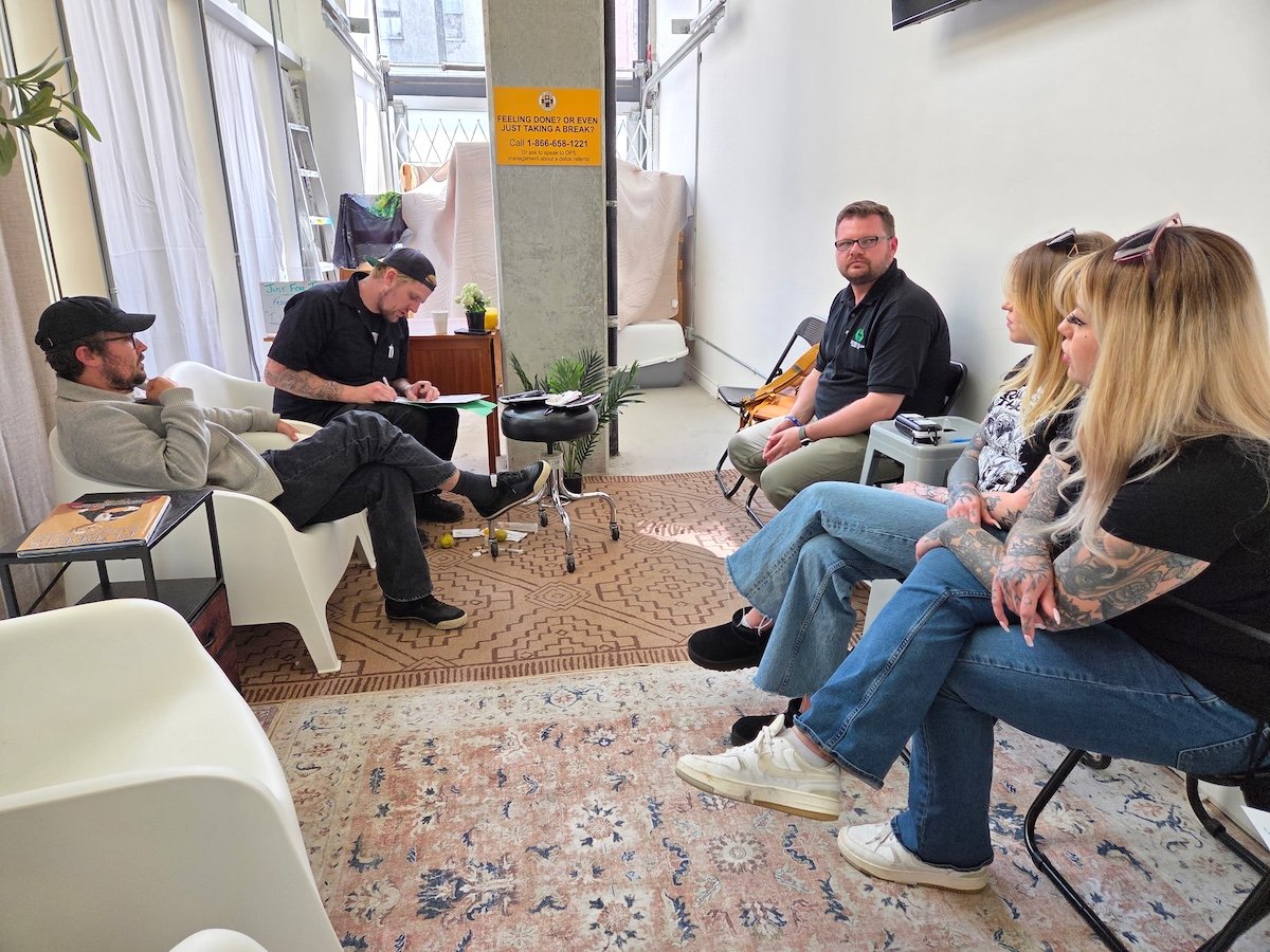 Five people sit on chairs in a semicircle. Trey Helten is taking notes. There are harm reduction supplies at his feet.