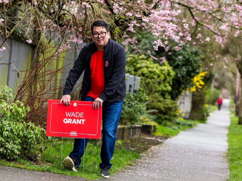 Wade Grant, a man in his mid-40s with medium-toned skin, puts an election sign up on a boulevard. He wears jeans, a red shirt and a black jacket.