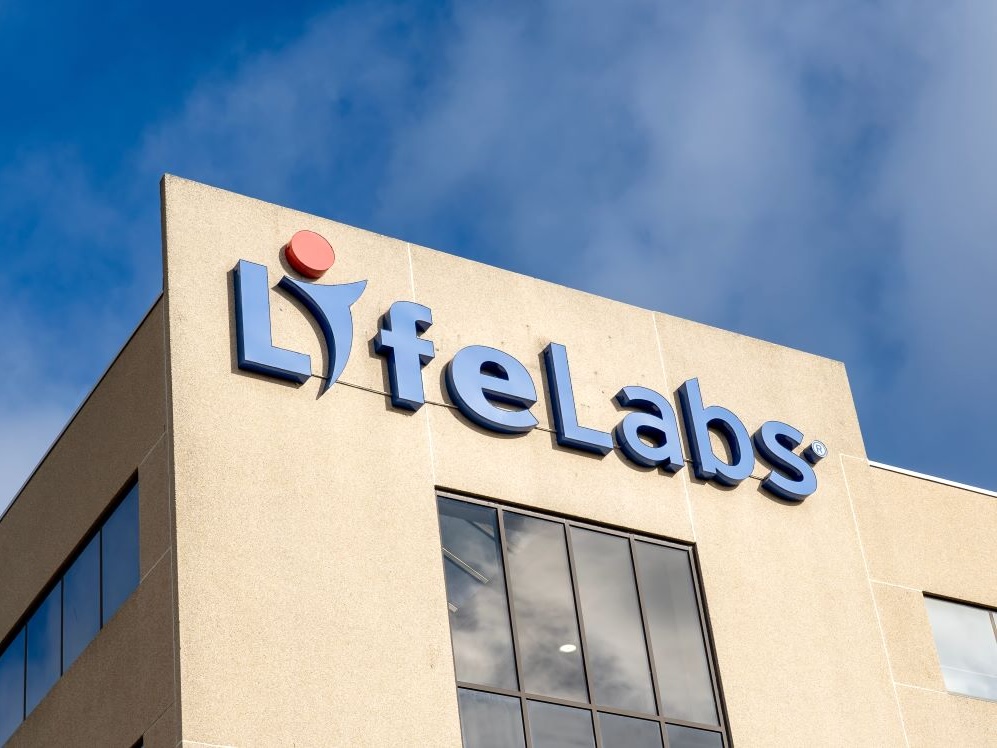 A sign on a beige office building reads ‘LifeLabs’ in blue lettering, with blue sky above.