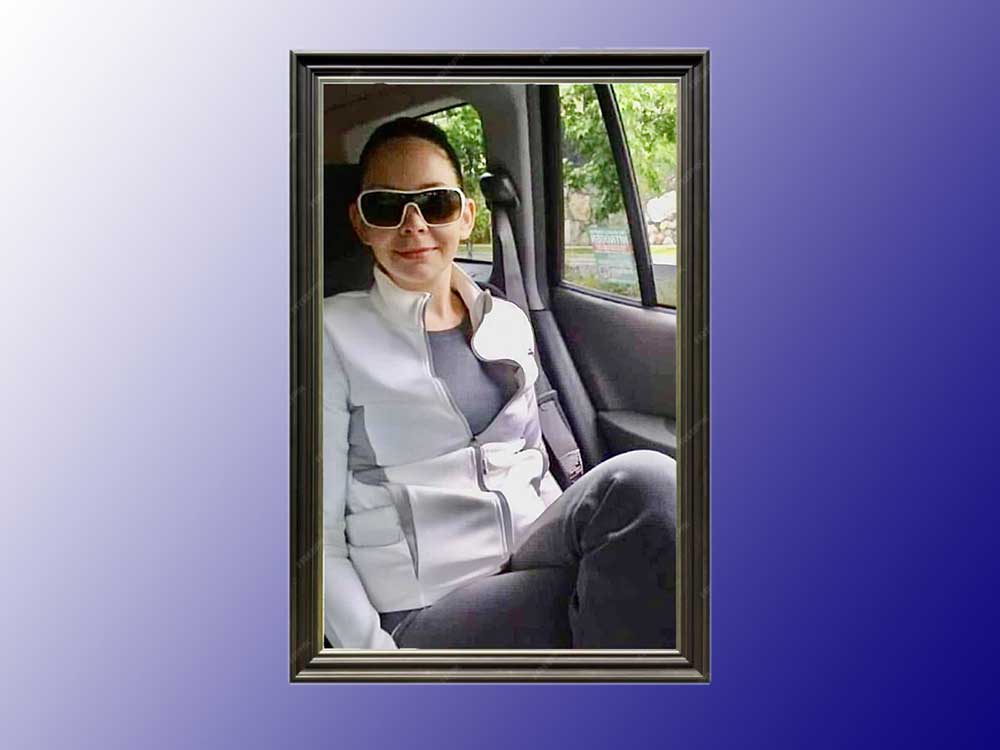 A young woman sits in a car and smiles at the camera. She wears a white jacket and sunglasses with white rims.