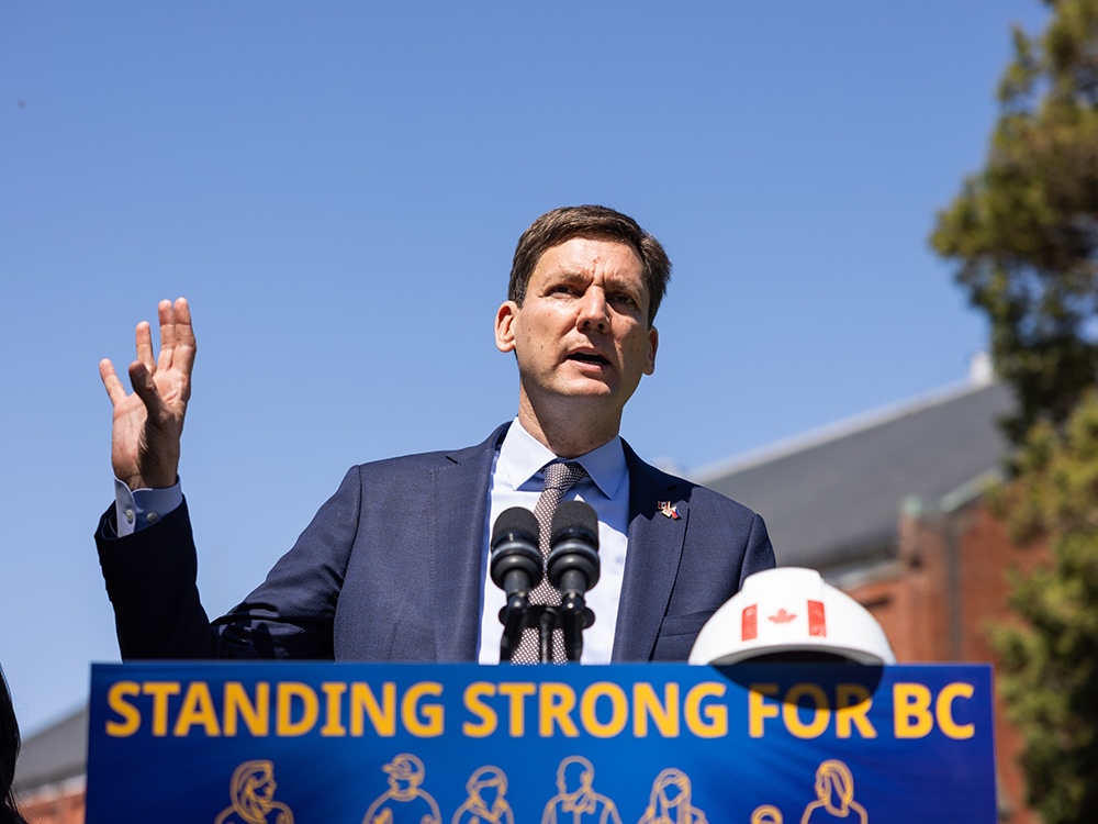 A man with light skin tone and short, brown hair stands at a podium and speaks into a microphone. There is a hard hat on the podium and blue sky in the background.