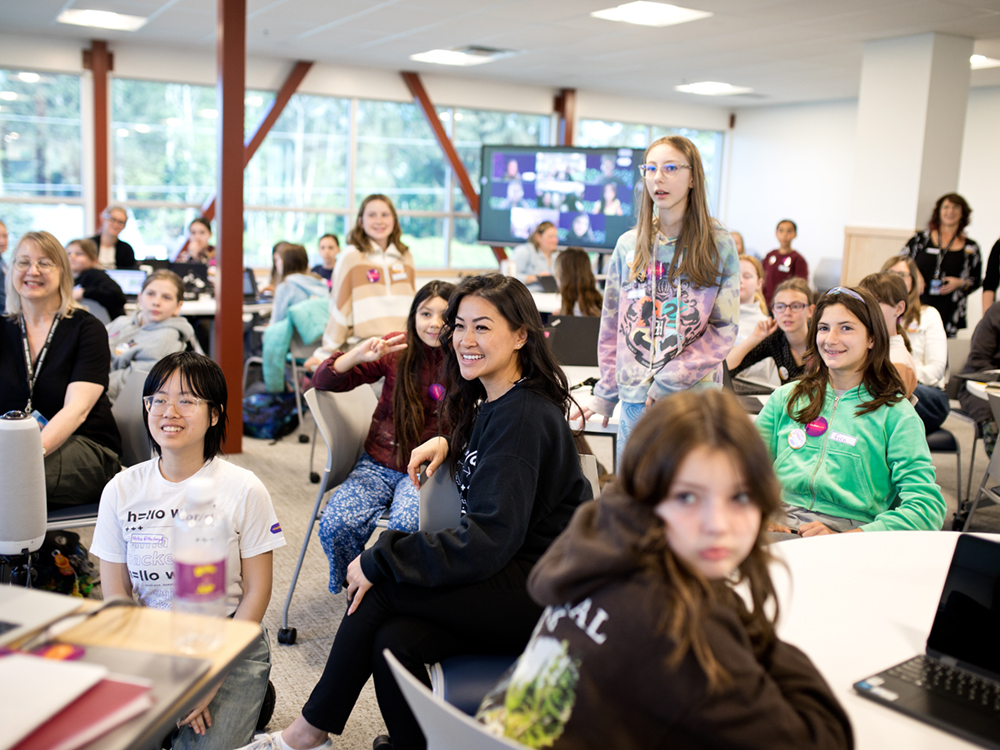 Approximately two dozen people, mostly children, face the camera as they sit at round tables in a brightly-lit meeting room.