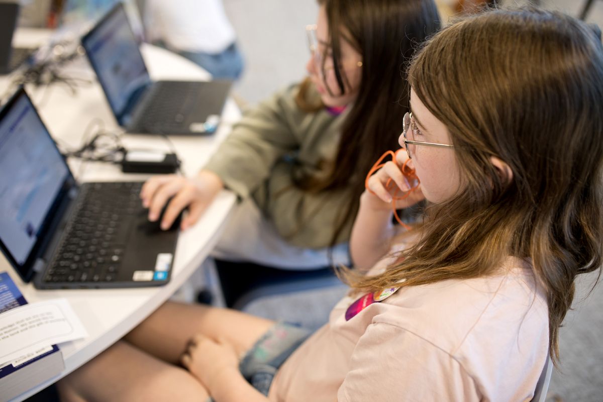 Looking over the shoulder at two light-skin toned children with long, straight brown hair and glasses, sitting at a table and working on laptops.