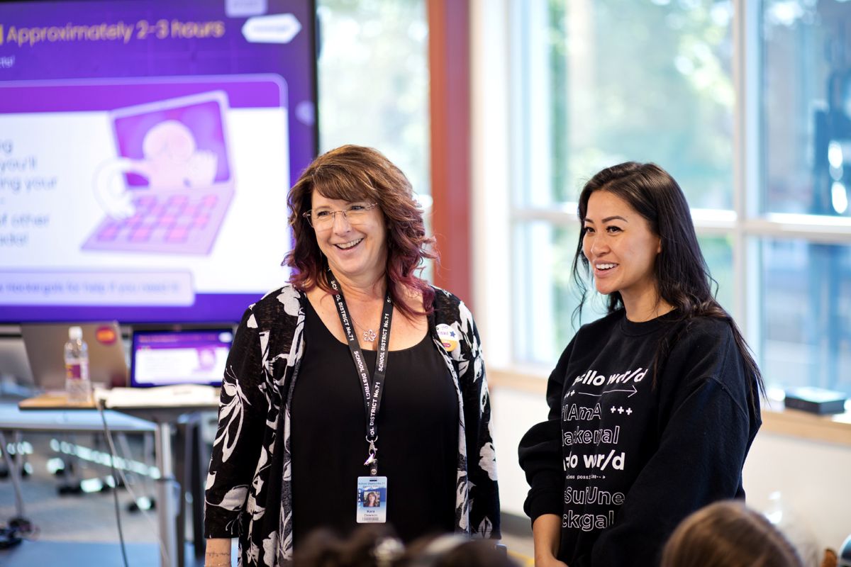 A woman with light skin tone and brown, curly shoulder length hair and glasses stands next to a medium-skin tone woman with straight hair below her shoulders. Behind them to the left is a projector screen and to the right a window.