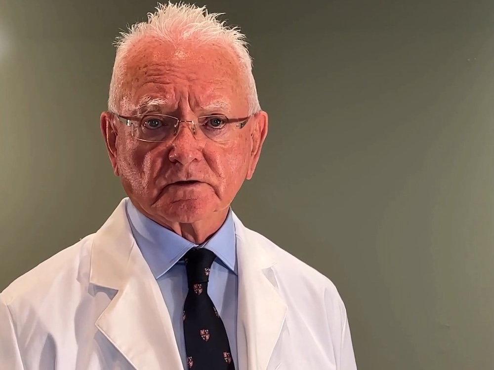 An 80-year-old man with light skin, short white hair and spectacles, wearing a white lab coat over shirt and tie.
