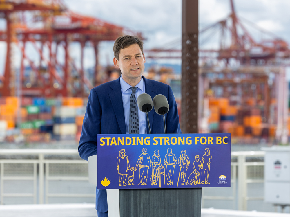 Premier David Eby, a middle-aged man with light skin tone and short brown hair, wears a blue suit and stands at a podium reading &lsquo;Standing Strong for BC.&rsquo;