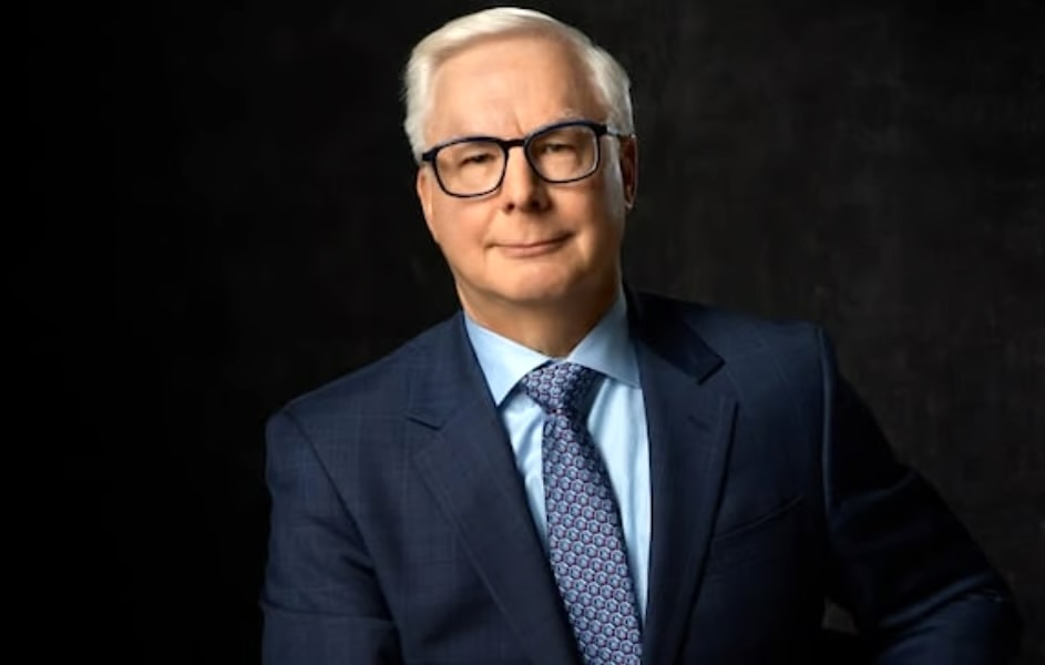 A man with light skin, short white hair and black-framed glasses wearing a navy blue suit.
