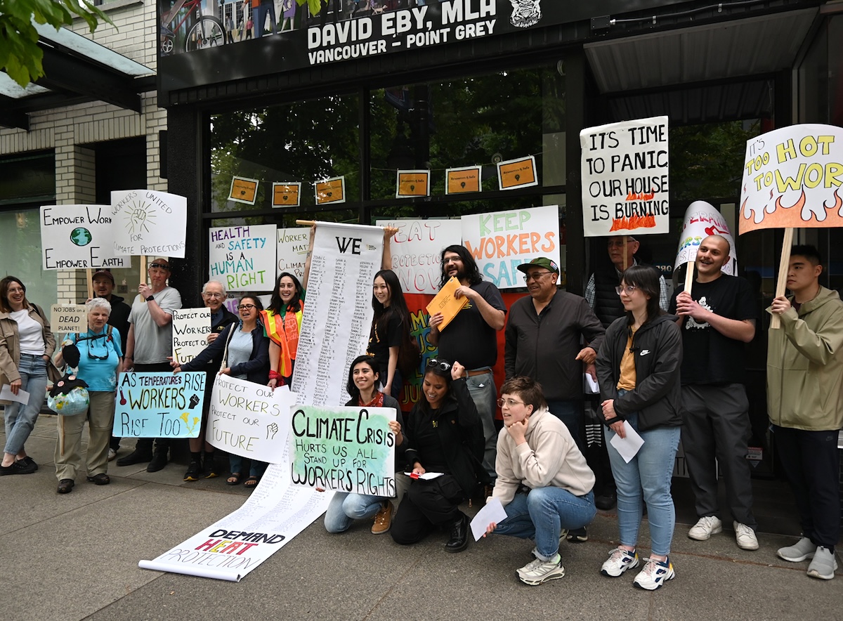A group of people stand with placards and an unfurled banner outside of David Eby’s MLA office. 