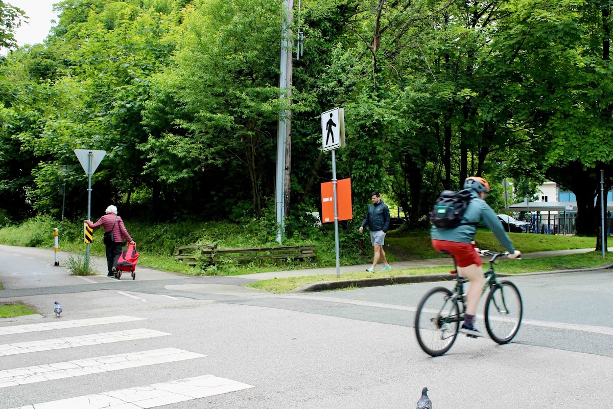 A cyclist and pedestrians move in front of a treed green space by an urban trail.