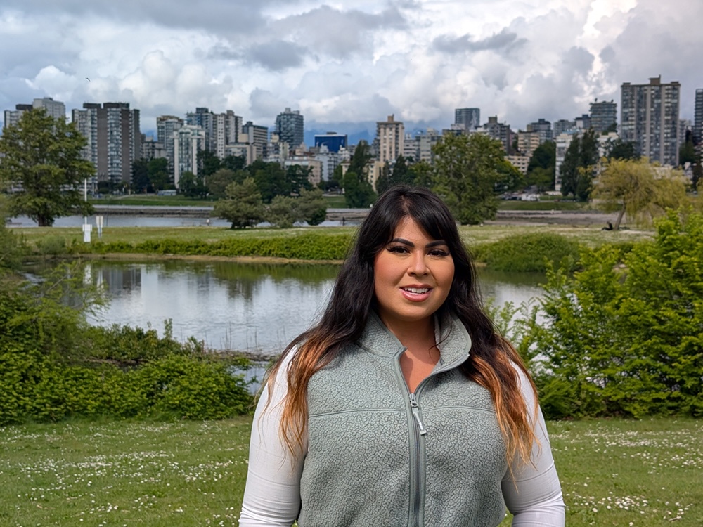 A person with medium skin tone and long, dark, ombre balayaged hair stands in a park with bodies of water. In the background is a city skyline.