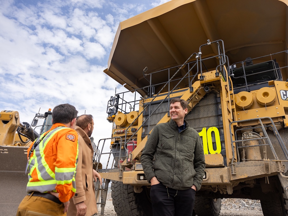 Premier David Eby, a tall, light-skinned 48-year-old man, smiles and speaks to two people in front of a giant yellow mining truck.