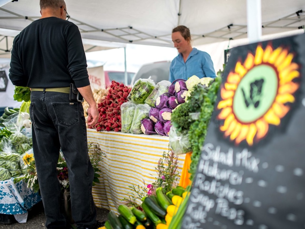 A person stands in front of a display of produce including radishes, salad mix and kohlrabi. They are being served by a person behind the display.