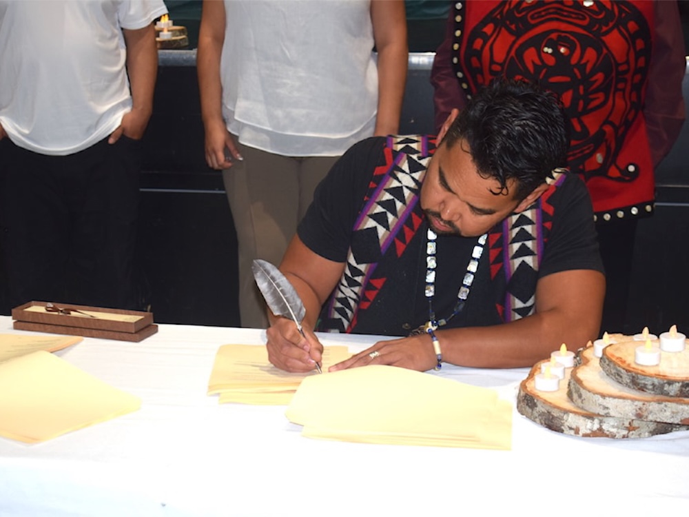 A man with dark wavy hair and medium skin tone wearing a red and black ceremonial vest is seated at a white table signing a set of yellow papers with a feathered pen. Three people are standing behind him.