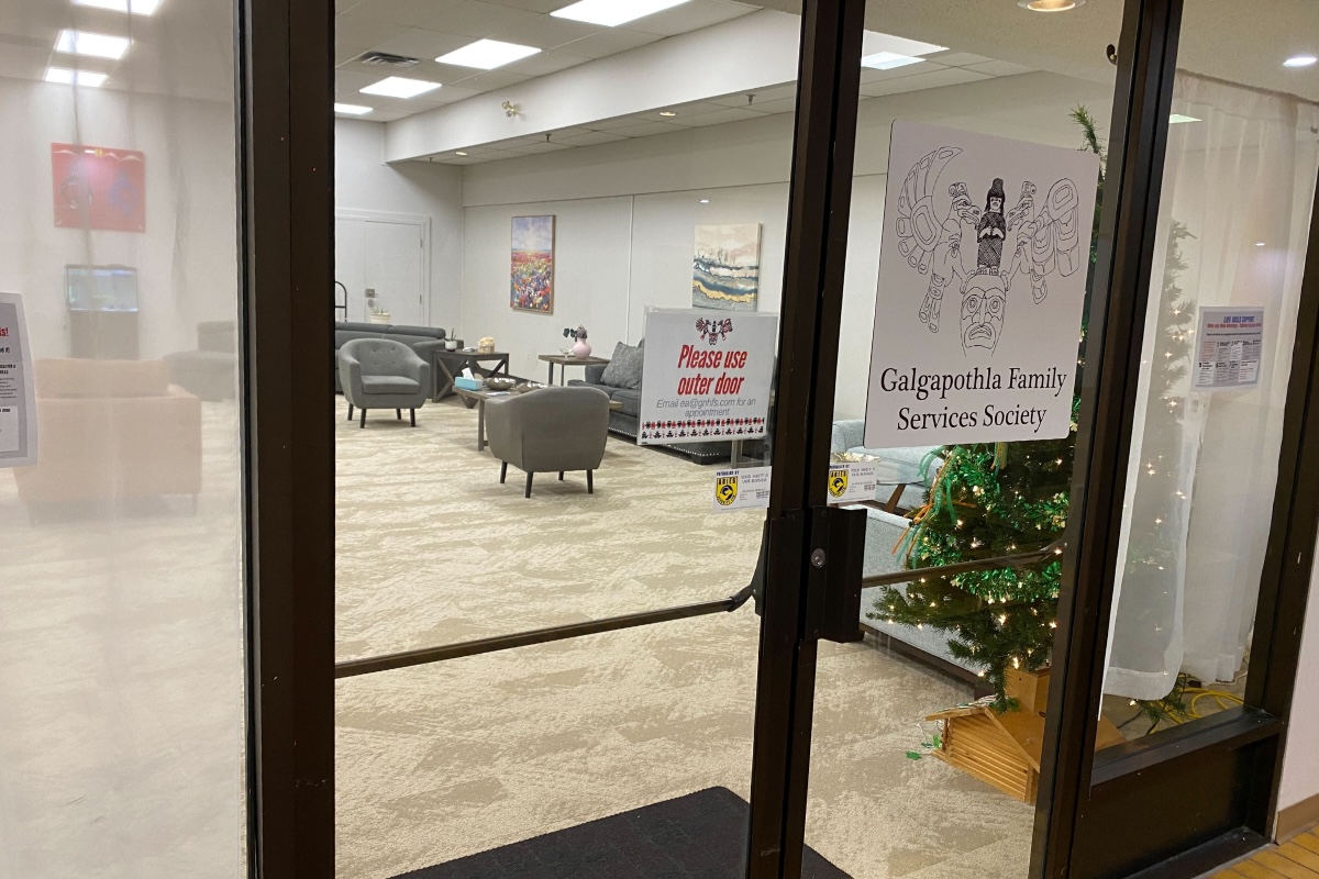 A clear door with black trim features a white sign that says &lsquo;Galgapothla Family Services Society&rsquo; under a black and white illustration. Inside is a seating area.