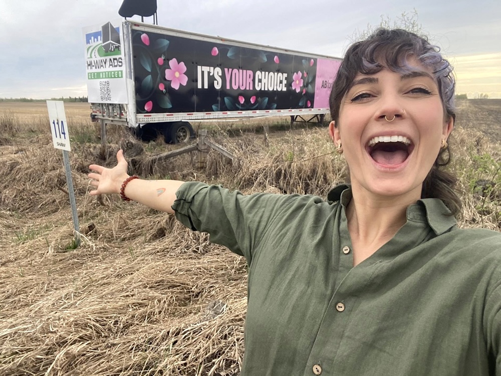 A woman with dark wavy brown and lilac hair and a green shirt stands in front of a white truck parked in a grassy field. Her arm is flung open and she looks overjoyed. ‘It’s your choice’ is written in large pink and white letters on the side of the truck.