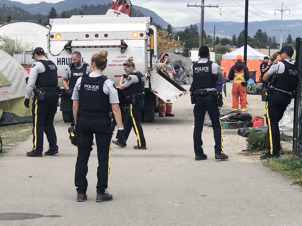 About eight RCMP officers wearing bulletproof vests gather around four or five city workers and a dump truck. Tents and other camping gear can be seen both set up and gathered in piles.