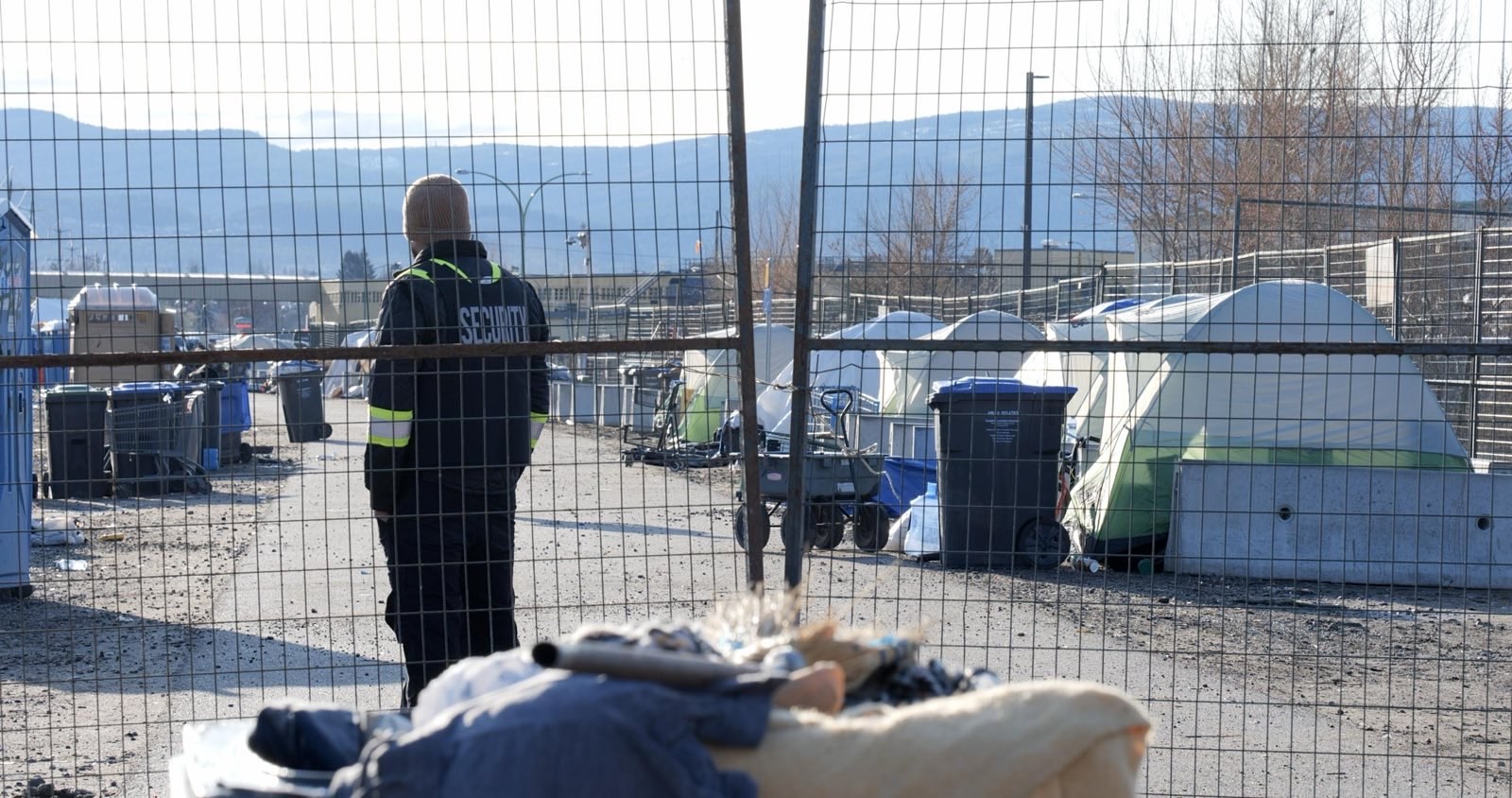 A security guard stands behind a gate. Tents are visible in the middle ground and background.