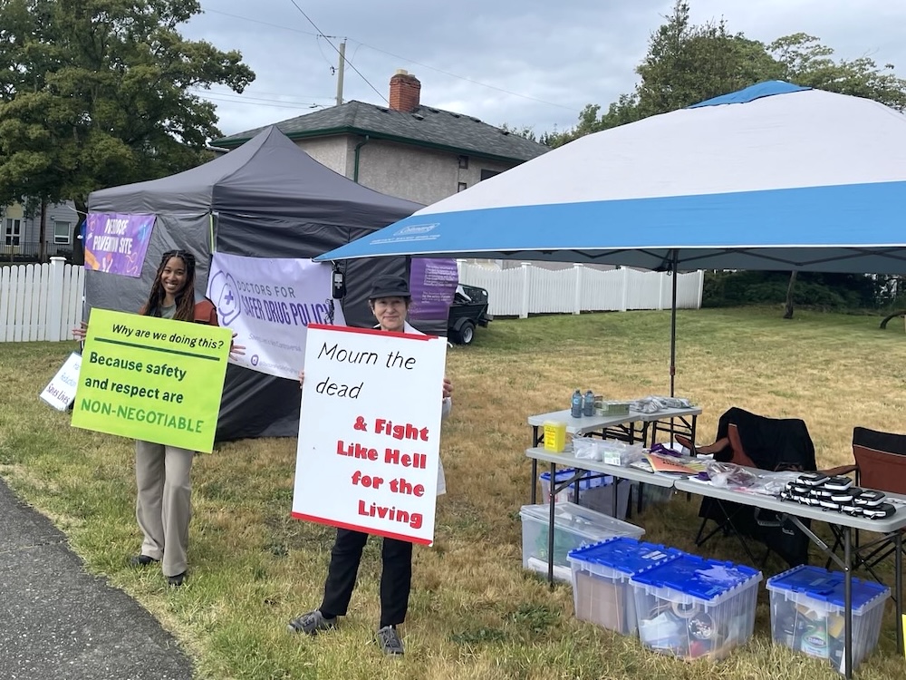 Two people stand on the grass beside a sidewalk, holding signs that read ‘Why are we doing this? Because safety and respect are non-negotiable’ and ‘Mourn the dead and fight like hell for the living.’ They’re standing in front of two tents and purple signs that read ‘Doctors for Safer Drug Policy.’ The blue and grey tent has two tables with harm reduction supplies neatly organized on it, and plastic boxes with additional supplies below it.