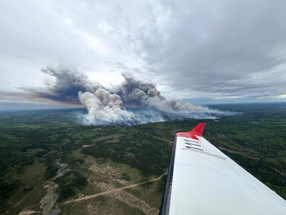 An aerial shot of two large plumes of grey smoke rising from a large area of forest. A plane’s wing is visible in the foreground.